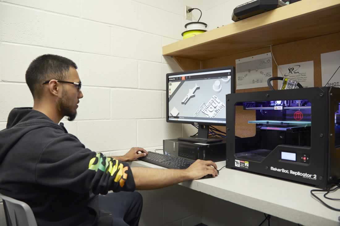 This is an image of a student working on a computer connected to a 3D printer.